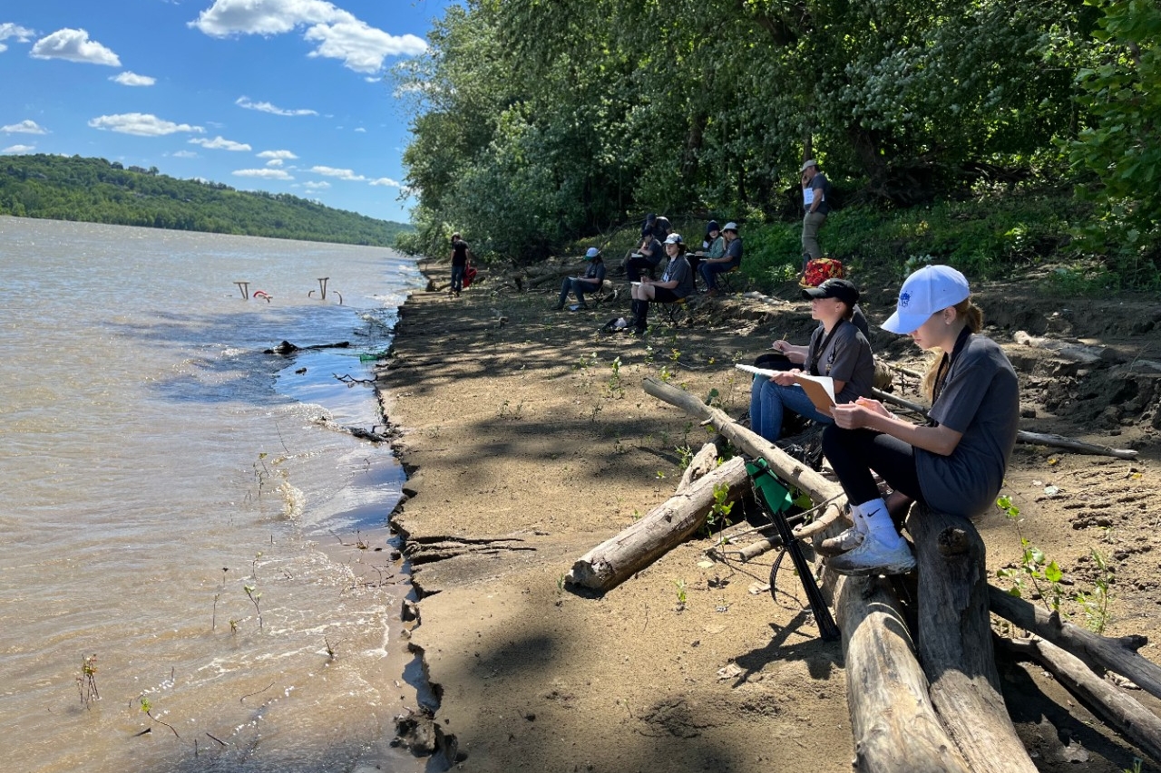 Middle school-aged kids sitting on driftwood along the shores of the Ohio River, drawing what they see in their art book.