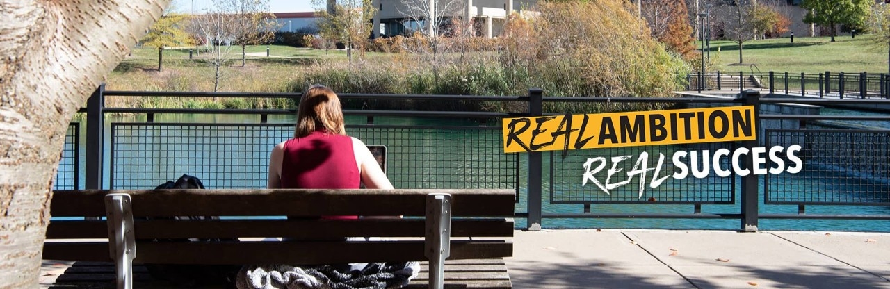 Student sitting on a bench by Loch Norse, working on laptop.