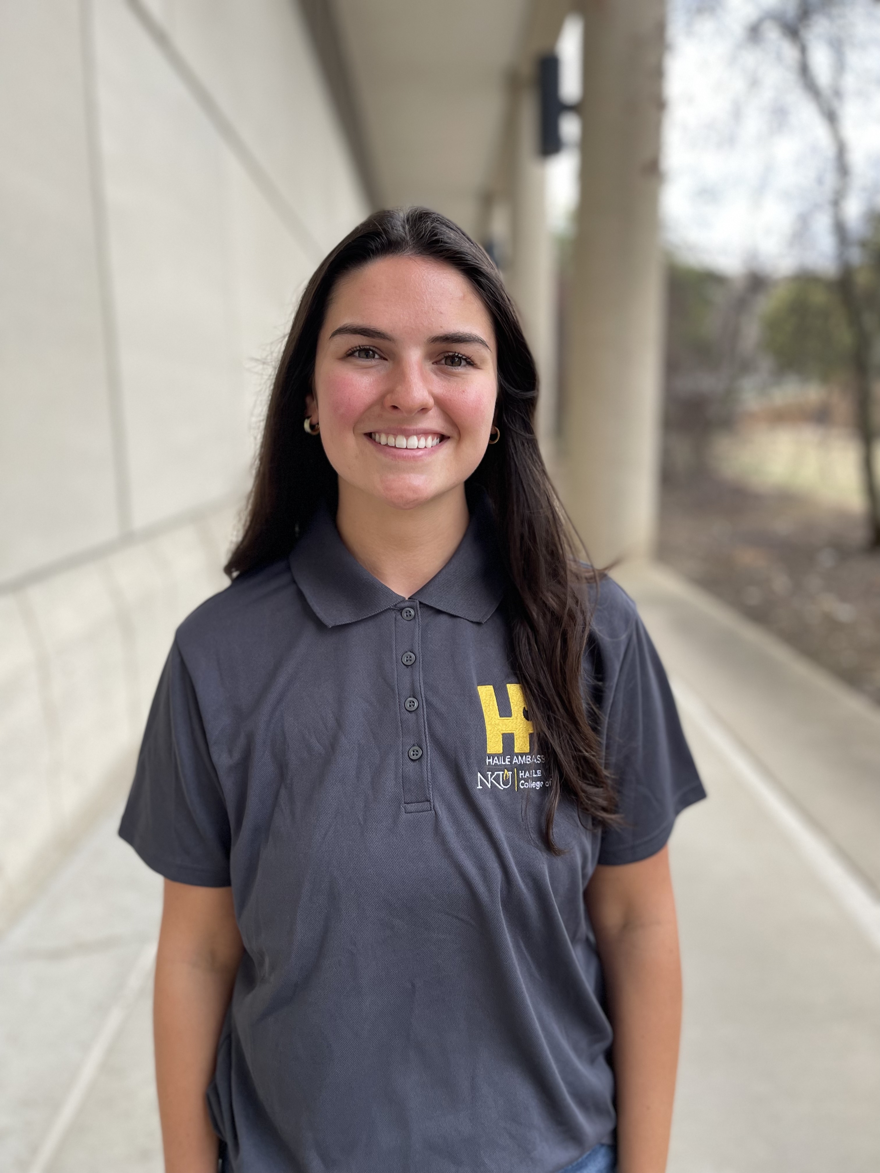 Samantha Deckard smiles while standing outdoors near a columned walkway. She has long dark hair and is wearing a gray NKU Haile Ambassadors polo shirt.