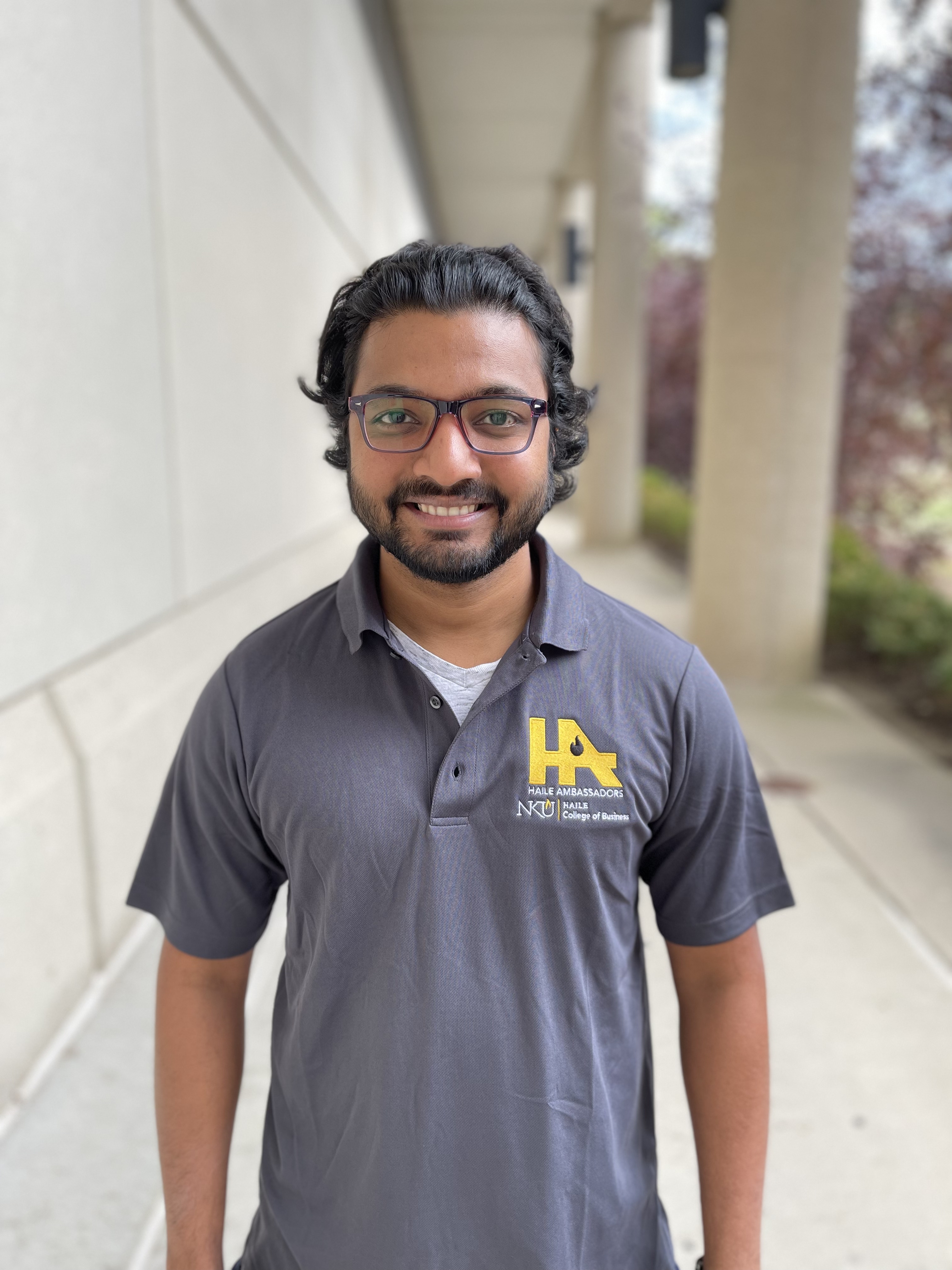 Jake Stokes smiles while standing outdoors near a columned building. He has dark hair, a beard, is wearing glasses, and a gray NKU Haile Ambassadors polo shirt.