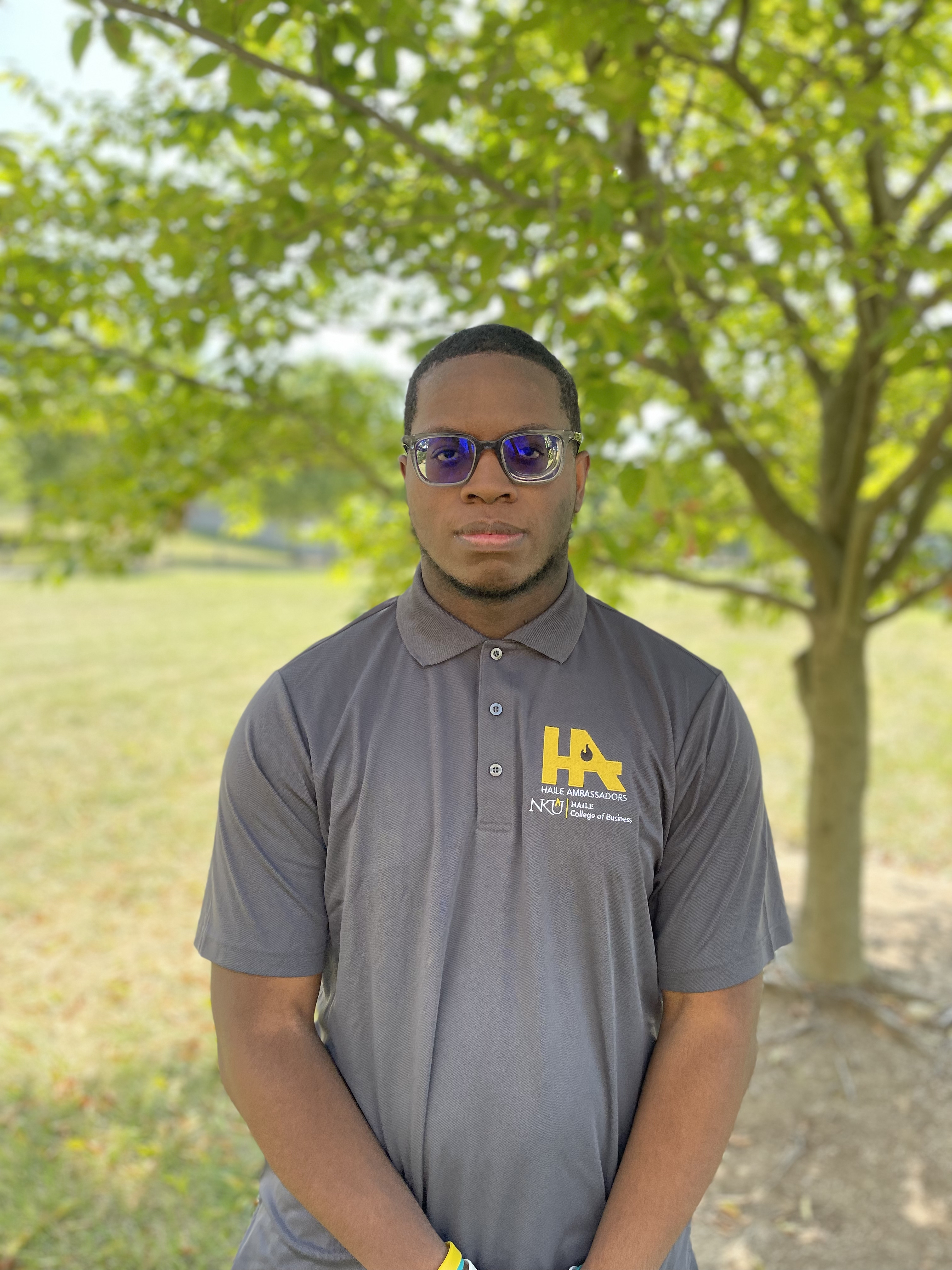 Taylor Shearer stands outdoors in front of a leafy tree and looks at the camera. He is wearing glasses and a gray NKU Haile Ambassadors polo shirt.
