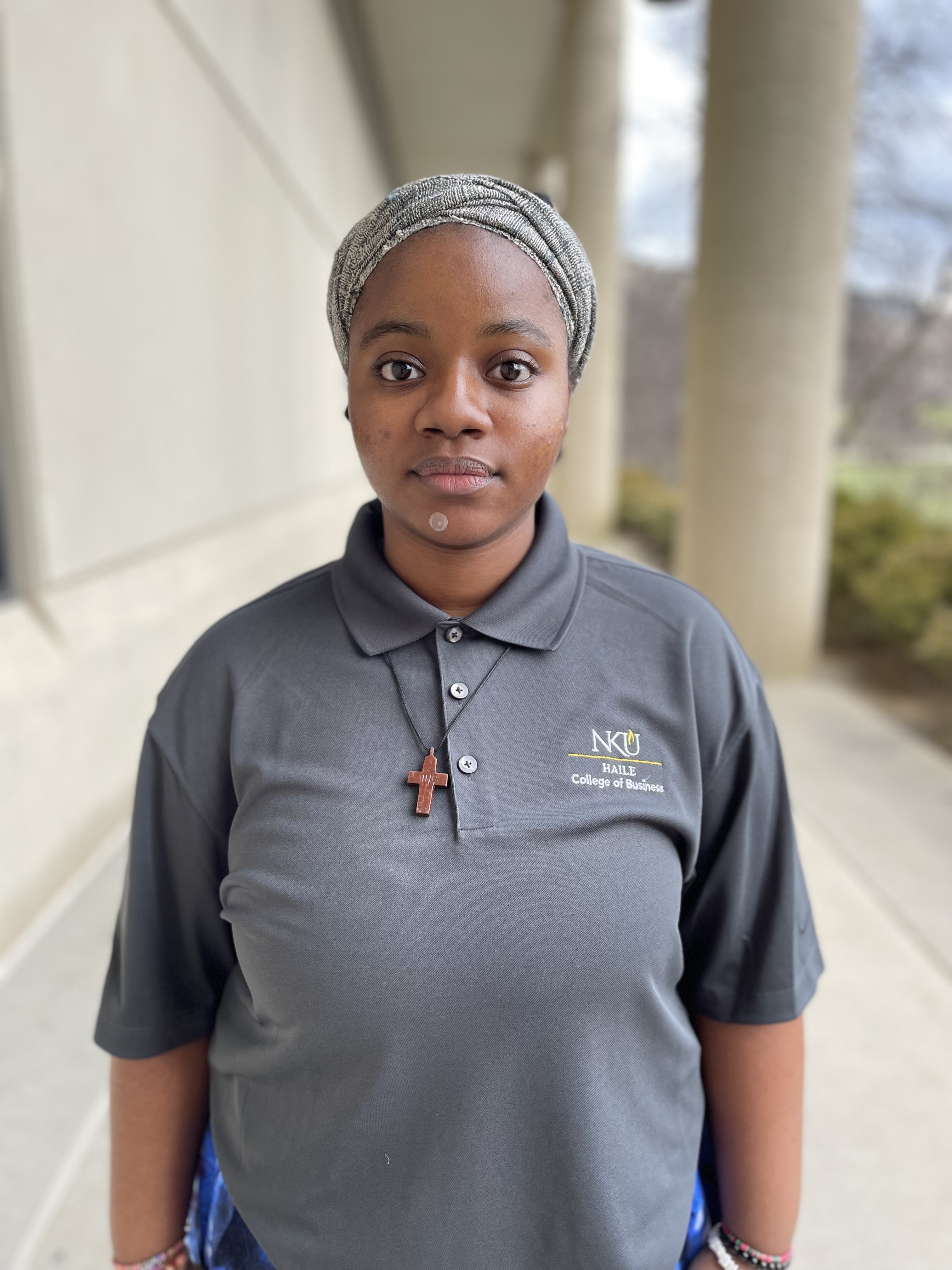 Princess Nworah stands outdoors near a columned building and looks at the camera. She is wearing a gray headwrap, a gray NKU Haile College of Business polo shirt, and a cross necklace.